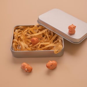 Könnte beinhalten: Eine rechteckige silberne Metalldose mit abnehmbarem Deckel, gefüllt mit hellbraunem Papierstreifen. Drei kleine, orangefarbene Capybara-Figuren sind zu sehen: eine in der Dose, eine auf dem Deckel und zwei vor dem Behälter platziert.