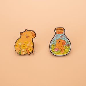 May include: Two enamel pins featuring a capybara. The first pin shows a capybara covered in yellow daisies. The second pin depicts a capybara inside a glass bottle with a cork stopper, surrounded by flowers, green foliage, and a small butterfly.