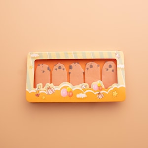 Op de afbeelding: Een rechthoekige doos met een helder venster toont vijf bruine, cartoonachtige, dierlijk gevormde plaknotities. De doos is oranje met gele en groene strepen en decoratieve illustraties van cadeaus, ballonnen en sterren.
