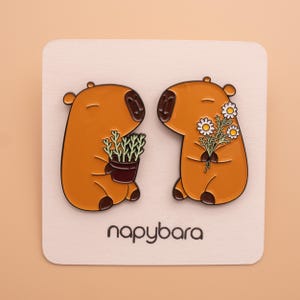 Peut inclure: Deux épingles en émail représentant des capybaras de dessin animé. L'une tient une plante en pot, l'autre un bouquet de marguerites. Les épingles sont marron avec des détails noirs et sont posées sur une carte blanche avec le mot "napybara" en noir.
