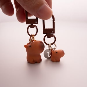 May include: Two brown capybara keychains with brown metal clasps and rings. One keychain has a small silver charm with the letter "L". The capybaras are small, cartoon-like figures.