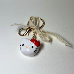 May include: A white Hello Kitty keyring with a red bow, black eyes, and whiskers. The keyring has a gold-coloured clasp and ring, and is adorned with a cream-coloured lace ribbon.