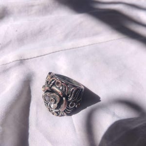 Peut inclure: Une bague argentée ornée d'un motif floral détaillé. La bague présente une rose proéminente au centre, entourée de motifs de feuilles et de vignes complexes. Des accents dorés sont visibles sur la surface de la bague, contrastant avec l'argent.
