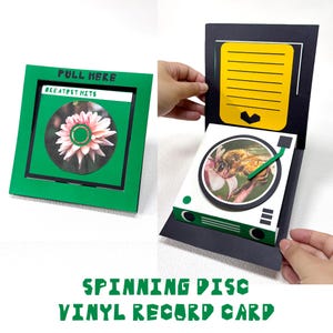 May include: A square green greeting card featuring a pull-tab mechanism labelled PULL HERE and GREATEST HITS. When opened, it reveals a pop-up design with a yellow lined writing area and a circular image of a bee on a white record player base.