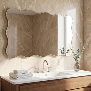 May include: A rectangular wall mirror with a wavy, organic-shaped gold frame. The mirror is mounted above a white bathroom sink vanity with a wooden cabinet, featuring a silver faucet, a stack of folded white towels, and a glass vase with greenery.