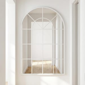 May include: White arched window-style mirror with a grid pattern. The mirror is mounted on a white wall in a hallway. The mirror reflects a room with hardwood floors and white walls. The mirror has the text "Casa Helena CH Mirrors".