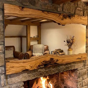 May include: A large, rustic wooden mirror is mounted on a stone fireplace. The mirror's frame is made of a thick, live-edge wood, and the fireplace has a fire burning in it. The room has a cozy, inviting atmosphere.