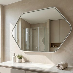 May include: A modern bathroom with a large, geometric mirror with a white frame. The mirror reflects a shower and neutral-toned walls. A countertop holds a soap dispenser, a small plant, and a rolled towel. The bathroom has a minimalist design.