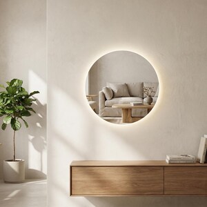 May include: A round, illuminated mirror hangs on a neutral-toned wall, reflecting a living room scene. Below, a wooden console holds books and vases. A potted plant and a wooden table are on the left. The mirror's diameter is approximately 36 inches.