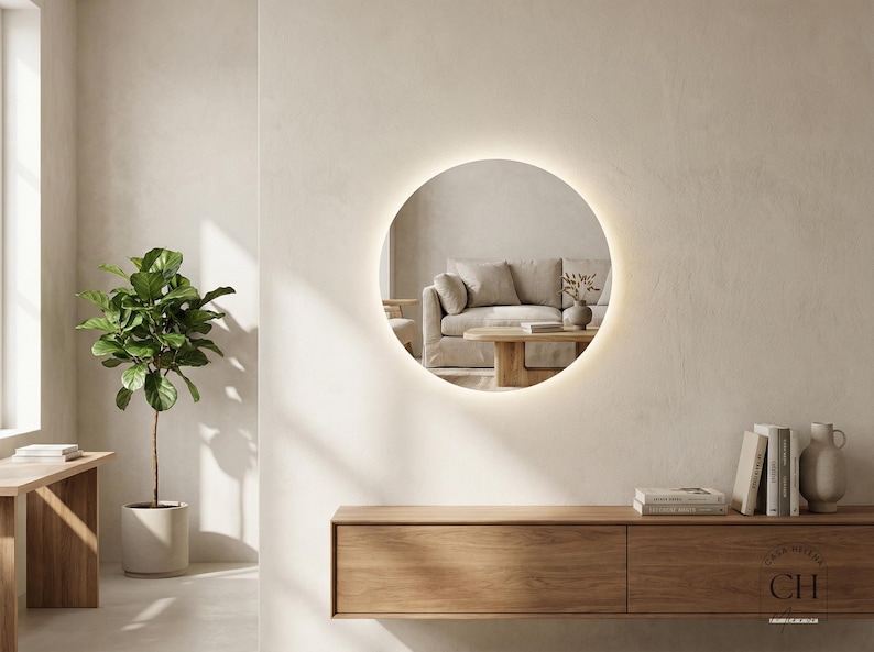 May include: A round, illuminated mirror hangs on a neutral-toned wall, reflecting a living room scene. Below, a wooden console holds books and vases. A potted plant and a wooden table are on the left. The mirror's diameter is approximately 36 inches.