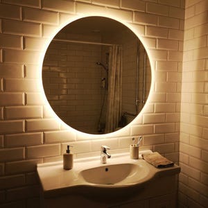 May include: A round, illuminated mirror is the focal point in this bathroom scene. The mirror is surrounded by a soft, glowing light and is mounted on a white brick-patterned wall. A white sink, faucet, and accessories complete the bathroom setup.