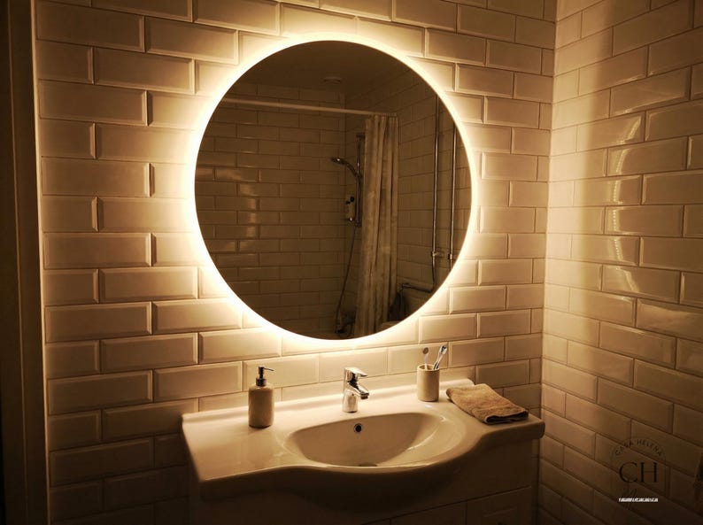 May include: A round, illuminated mirror is the focal point in this bathroom scene. The mirror is surrounded by a soft, glowing light and is mounted on a white brick-patterned wall. A white sink, faucet, and accessories complete the bathroom setup.