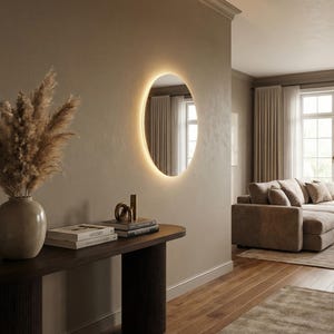 May include: An interior design scene featuring a modern, illuminated circular mirror on a neutral-toned wall. A dark wood console table holds books, decorative objects, and a vase with dried pampas grass. The room has a warm, inviting atmosphere.