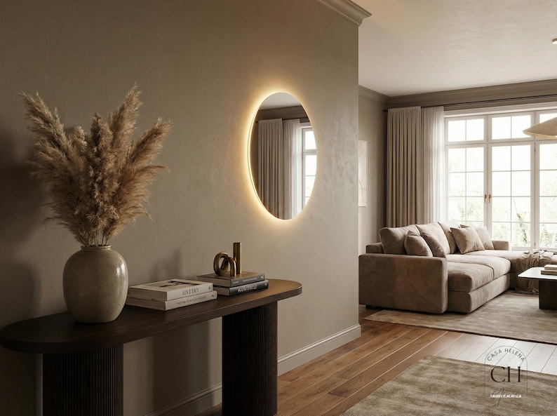 May include: An interior design scene featuring a modern, illuminated circular mirror on a neutral-toned wall. A dark wood console table holds books, decorative objects, and a vase with dried pampas grass. The room has a warm, inviting atmosphere.