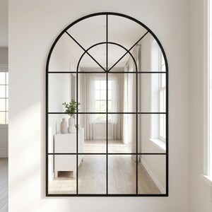 May include: Large arched mirror with a black grid design. The mirror reflects a bright interior with white walls, a white cabinet, and a window. The mirror is a decorative element for home decor.