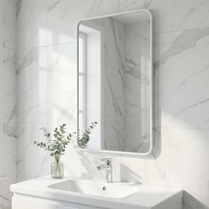 May include: A rectangular white-framed mirror hangs above a white sink in a bathroom. The mirror reflects a window and the marble-patterned wall. A small glass vase with green leaves sits on the counter. The wall is tiled with white marble.