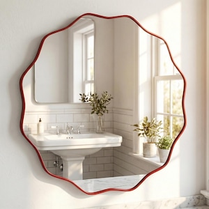 May include: A red, wavy-edged mirror hangs on a white wall above a bathroom sink. The mirror reflects the room, including a window and plants. The bathroom has white tile and a marble countertop. The mirror's frame is a vibrant red.