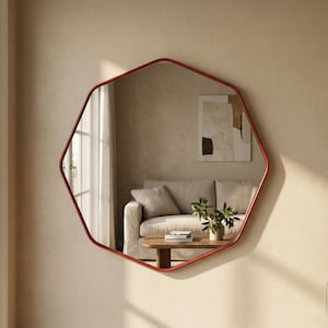 Op de afbeelding: Een rood omkaderde, geometrische spiegel hangt aan een beige muur. De spiegel weerspiegelt een woonkamerscène met een bank, salontafel en kunstwerken. De vorm van de spiegel is een onregelmatige achthoek.
