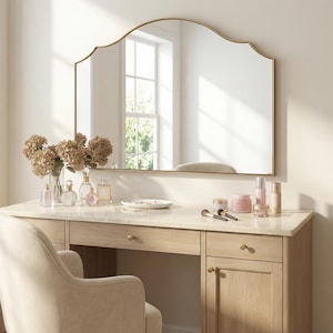 Könnte beinhalten: Ein Schminktisch-Set mit einem großen, bogenförmigen, goldgerahmten Spiegel. Der helle Holzschminktisch hat eine Marmorplatte mit Schönheitsprodukten und Make-up-Pinseln. Ein beige gepolsterter Stuhl steht davor. Eine Vase mit getrockneten Blumen steht auf dem Tisch.