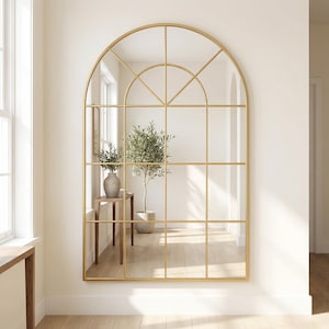 Op de afbeelding: Een grote, boogvormige spiegel met een gouden metalen frame, ontworpen als een raam. De spiegel is verdeeld in rechthoekige en boogvormige panelen. Hij hangt aan een witte muur in een kamer met een houten vloer en planten.