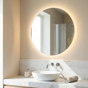 May include: A round, backlit mirror is the focal point in this bathroom scene. Below the mirror is a white sink, a faucet, and a rolled towel. The countertop is marble, and the walls are a neutral color.