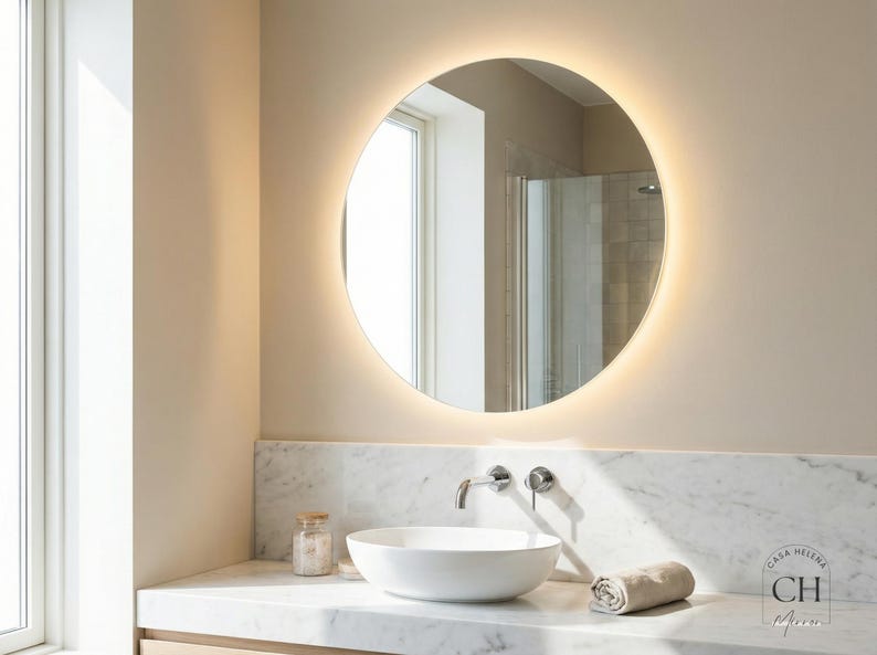 May include: A round, backlit mirror is the focal point in this bathroom scene. Below the mirror is a white sink, a faucet, and a rolled towel. The countertop is marble, and the walls are a neutral color.