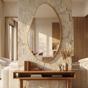 Peut inclure: Un miroir mural de forme organique irrégulière avec un cadre doré fin, fixé sur un mur en pierre texturée. Sous le miroir se trouve une console en bois rectangulaire portant une petite sculpture en céramique, un vase d'herbes séchées et des livres.