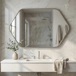 May include: A modern bathroom with a large, hexagonal mirror with a brushed metal frame. The mirror reflects a shower and towels. A white countertop holds a sink, faucet, and a vase with greenery. A folded towel rests on the counter.