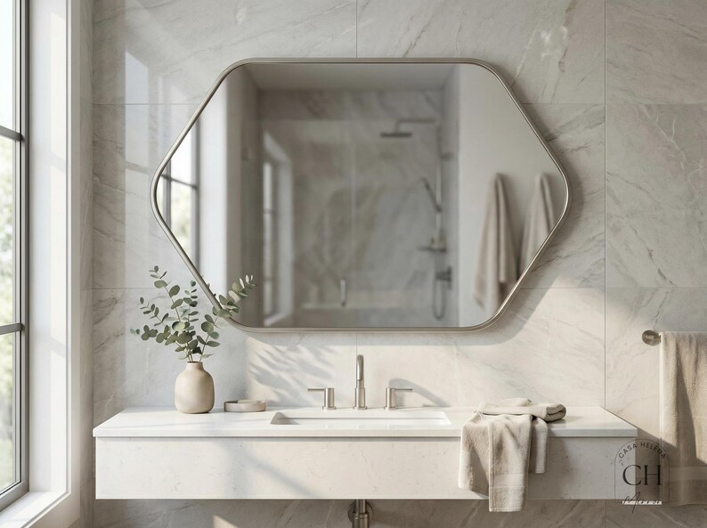 May include: A modern bathroom with a large, hexagonal mirror with a brushed metal frame. The mirror reflects a shower and towels. A white countertop holds a sink, faucet, and a vase with greenery. A folded towel rests on the counter.