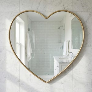 May include: A heart-shaped mirror with a gold frame hangs on a white marble tiled wall. The mirror reflects a bathroom with a shower, sink, and white towels. The mirror is a decorative accent.