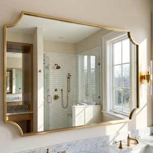 以下が含まれることがあります： 装飾的な上端を持つ長方形の金色のフレームの鏡が、大理石のカウンタートップの上に吊り下げられています。 透明なガラス管が付いた2つの金の燭台が鏡の両側にあります。 鏡はシャワーと窓のあるバスルームを映しています。