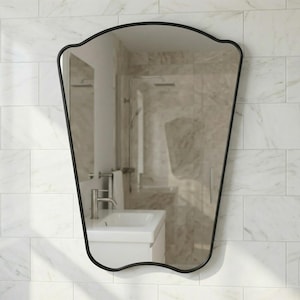 May include: A black-framed wall mirror with a unique, curved top and a tapered shape. The mirror reflects a bathroom interior with a white sink and marble-like tile. The mirror is mounted on a white tiled wall.