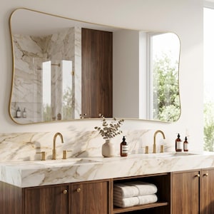 May include: A large wall mirror with an irregular, organic wavy shape and a thin gold metal frame. The mirror is mounted above a double vanity with a white marble countertop, gold taps, and dark wood cabinetry with open shelving.