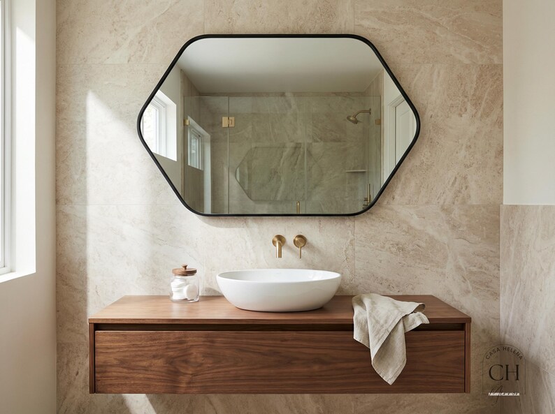 May include: A modern bathroom featuring a hexagonal black-framed mirror above a floating wooden vanity. The vanity holds a white vessel sink, gold fixtures, and a linen towel. The walls are tiled in a neutral stone pattern.