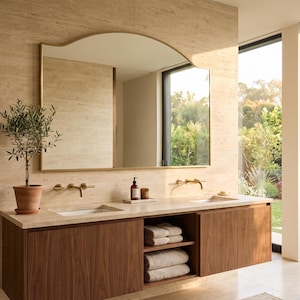 May include: A double vanity bathroom featuring a wooden cabinet with two sinks, a large arched mirror with a thin gold frame, and a potted plant. The vanity includes open shelving stacked with folded towels and closed storage cabinets.