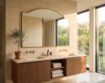 May include: A double vanity bathroom featuring a wooden cabinet with two sinks, a large arched mirror with a thin gold frame, and a potted plant. The vanity includes open shelving stacked with folded towels and closed storage cabinets.