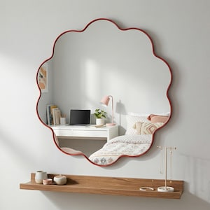 Op de afbeelding: Een grote, decoratieve spiegel met een rood geschulpte lijst hangt aan een witte muur. Daaronder staat een houten plank met decoratieve items. De spiegel weerspiegelt een bureau met een laptop, een bed en een roze lamp.
