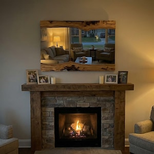 May include: A large rectangular mirror with a live edge wooden frame hangs above a stone fireplace. The mirror reflects a living room scene. The fireplace has a lit fire, and the room is lit by lamps.