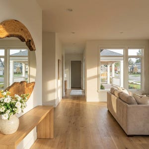 Pu&ograve; includere: Un interno moderno con un grande specchio ovale con finiture in legno naturale. Un tavolo consolle in legno ospita un vaso di fiori bianchi. La stanza ha pavimenti in legno e una palette di colori neutri, con vista sull'esterno attraverso ampie finestre.