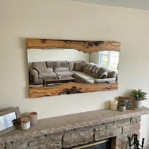 May include: A large rectangular mirror with a live edge wooden frame. The mirror reflects a living room with a sectional sofa. The frame is a natural wood colour with dark brown accents.