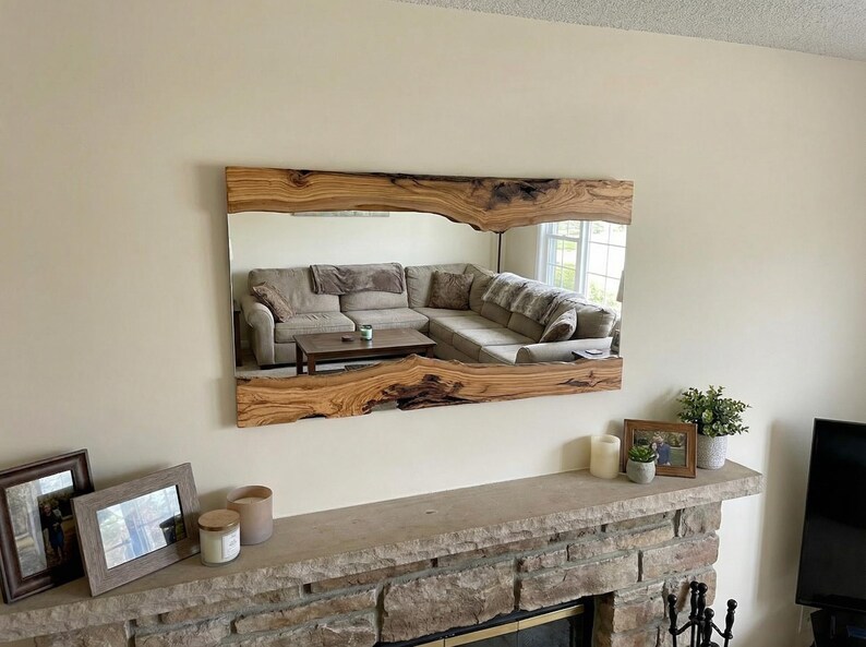 May include: A large rectangular mirror with a live edge wooden frame. The mirror reflects a living room with a sectional sofa. The frame is a natural wood colour with dark brown accents.