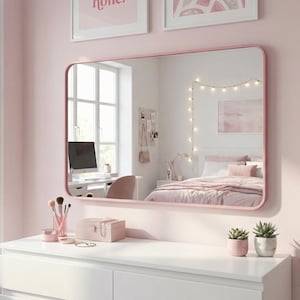 May include: A pink-framed mirror hangs above a white dresser, reflecting a bedroom scene. The room is decorated in shades of pink, with framed art and string lights. Makeup brushes, a jewelry box, and small potted plants sit on the dresser.