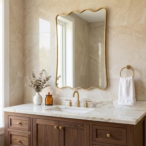 Puede incluir: Un mueble de baño con espejo con marco dorado, encimera de mármol y mueble de madera oscura. Una toalla blanca cuelga de un anillo dorado. Un jarrón blanco y un tarro marrón están sobre la encimera.