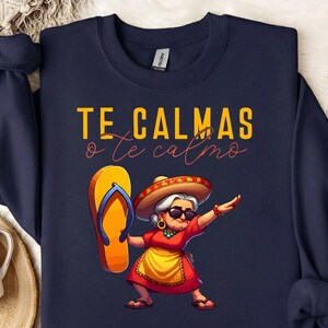 以下が含まれることがあります： 紺色のスウェットシャツに、黄色と赤で「TE CALMAS o te calmo」と書かれています。グラフィックは、ソンブレロ、サングラス、ビーチサンダルを持ったおばあさんの漫画です。女性は赤いドレスと黄色のエプロンを着ています。