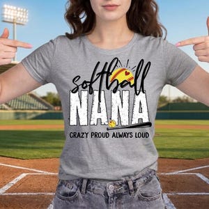 May include: A gray t-shirt with the words "Softball Nana" in a distressed white font, featuring a softball graphic with a heart. The text "Crazy Proud Always Loud" is below the main text. The background is a baseball field.