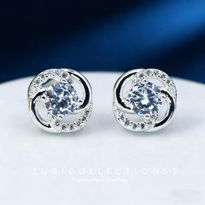 May include: A pair of silver stud earrings with a central clear gemstone surrounded by smaller clear stones. The earrings have a swirling design and are set against a white and blue background. The text "ZUBICOLLECTIONSS Handcrafted Jewellery" is at the bottom.