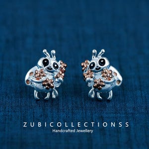 May include: A pair of silver-toned bee-shaped earrings with black eyes and copper-colored flower accents. The earrings are set against a dark blue background. The text "ZUBI COLLECTIONS S S Handcrafted Jewellery" is at the bottom.