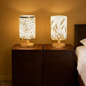 May include: Two decorative bedside lamps with wooden bases and cylindrical shades. One shade features a floral pattern, the other a wheat design. Both lamps sit on dark brown nightstands, with a bed visible in the background.