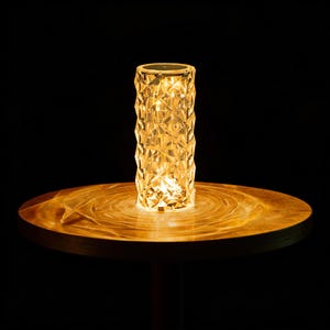 Op de afbeelding: Een cilindervormige kristallen tafellamp met een facetontwerp, die een warme, gouden gloed uitstraalt. De lamp staat op een houten tafel en creëert een lichtpatroon. De lamp is ongeveer 25 cm hoog.