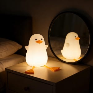 May include: A white, duck-shaped night light with orange beak and feet sits on a nightstand. The lamp is reflected in a round mirror. The lamp has small arms and is lit from within, creating a soft glow. The background is a dimly lit bedroom.